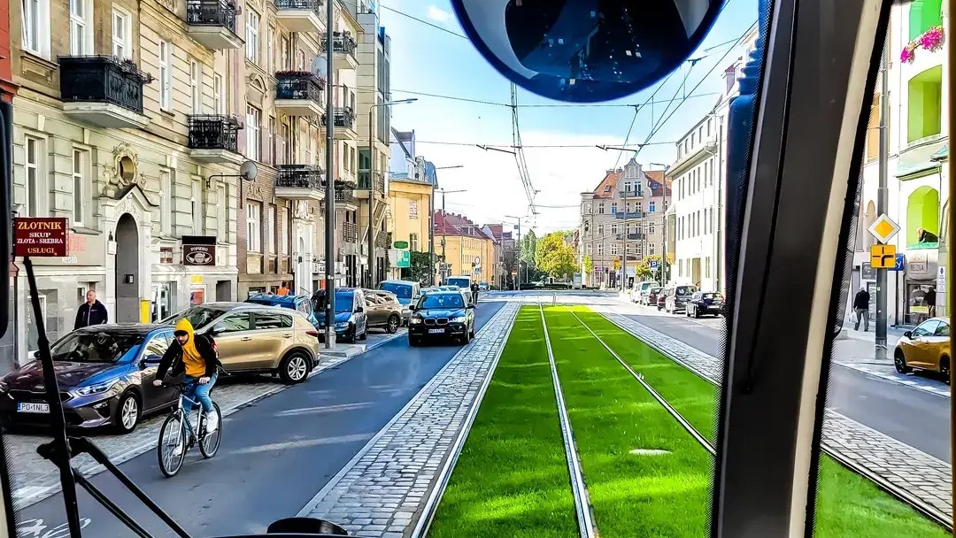 Ścieżką rowerową jedzie rowerzysta w żółtej bluzie. Po obu stronach ulicy stoją budynki mieszkalne i usługowe. Jest słoneczny dzień. Jezdnia i torowisko prowadzą w kierunku skrzyżowania widocznego w oddali.