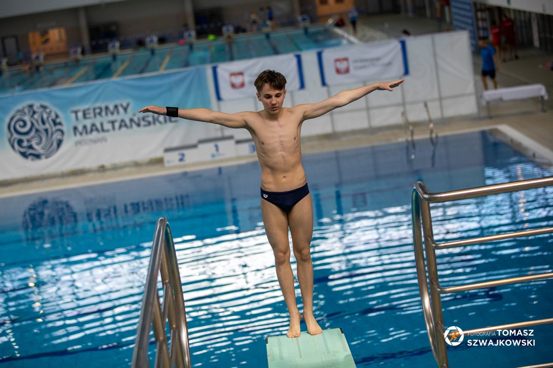 Skoki do wody w ramach Ogólnopolskiej Olimpiady Młodzieży w sportach letnich, fot. Tomasz Szwajkowski - mężczyzna stoi tyłem na pomoście przed skokiem do wody na pływalni. W tle widać pływalnię