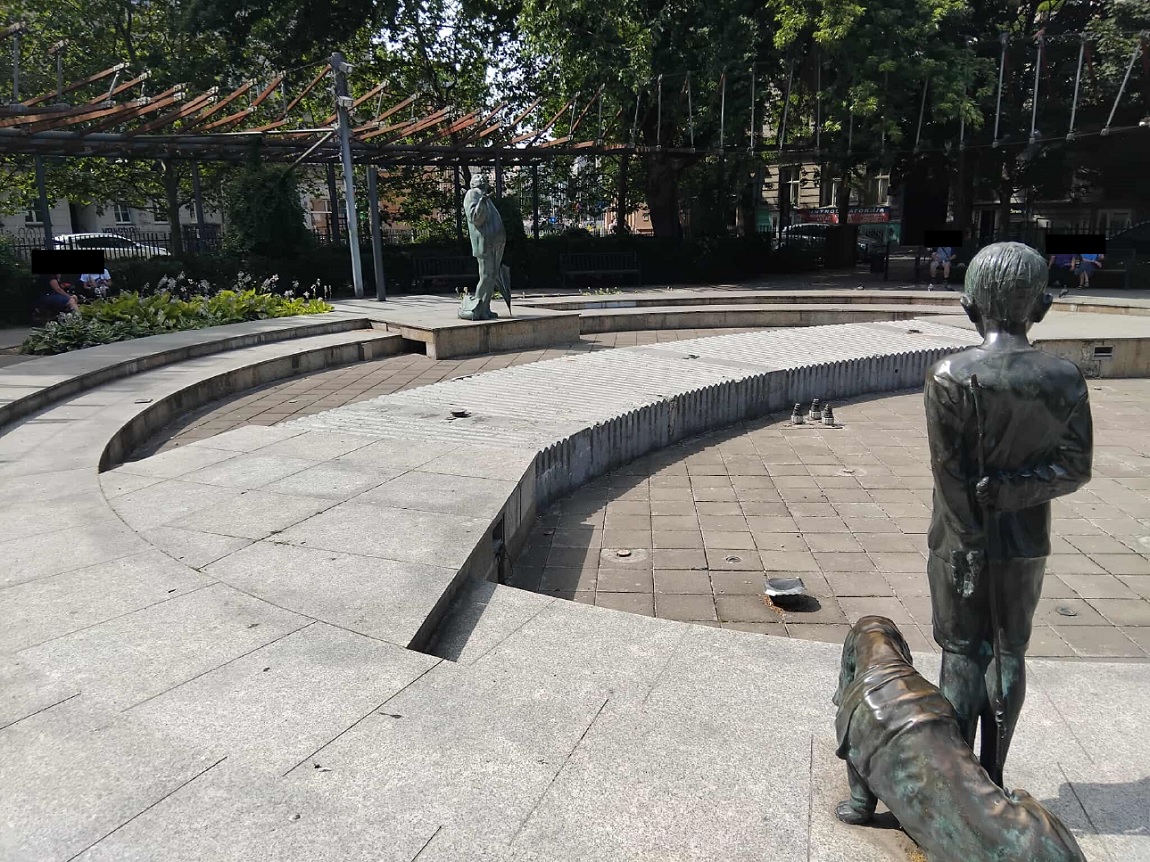 skwer otoczony zielenią, niedziałąjąca fontanna, jej elemantami są rzeźby starszego mężczyzny i chłpca z psem