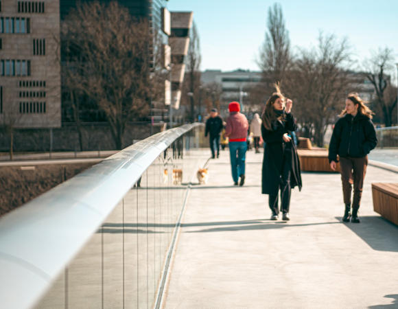 Słoneczny, betonowy chodnik z metalową barierką po lewej i drewnianą ławką po prawej. Na pierwszym planie idą dwie uśmiechnięte młode kobiety w ciemnych kurtkach.