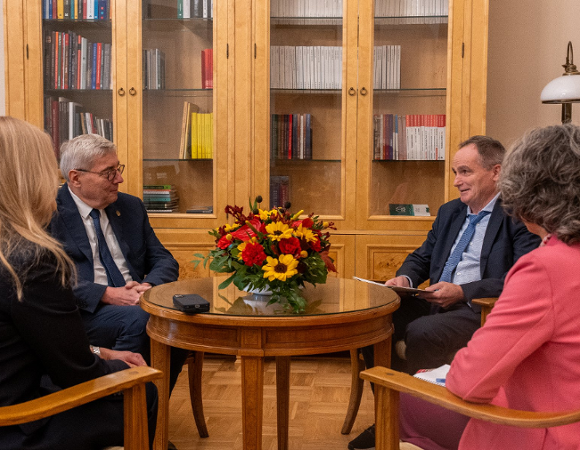 Spotkanie czterech osób przy okrągłym stole z bukietem kwiatów w gabinecie z drewnianą biblioteczką w tle.
