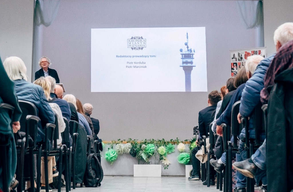 spotkanie w dużej sali ludzie siedzą na krzesłach ustawionych w rzędach na scenie jedna osoba stoi przy mówinicy za nią wyświetlony ekran z prezentacją