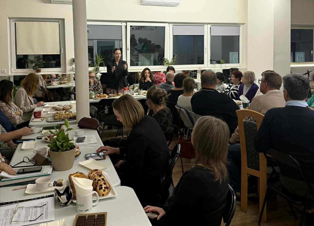 spotkanie w osiedlowej świetlicy przy zastawionych stołach wiele osób w różnym wieku jedna osoba kobie stoi i przemawia do pozostałych