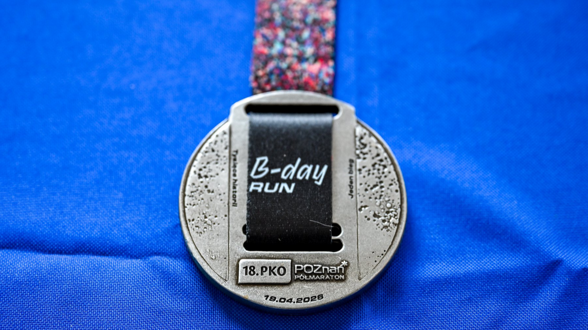 Srebrny, okrągły medal pamiątkowy z 18. PKO Poznań Półmaratonu, leżący na niebieskim materiale. Na medalu widnieje czarna wstążka z białym napisem "B-day run". W dolnej części wygrawerowano nazwę biegu oraz datę 19 kwietnia 2026. Medal ma surową, lekko porowatą teksturę po bokach, a u góry przymocowana jest kolorowa, wzorzysta taśma.