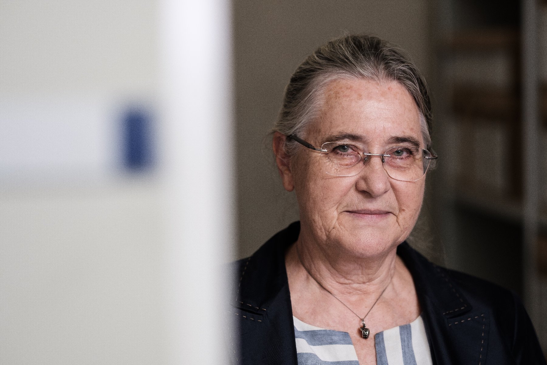 Starsza kobieta w okularach bez oprawek i marynarce. Uśmicha się delikatnie. Ma łańcuszek na szyi.