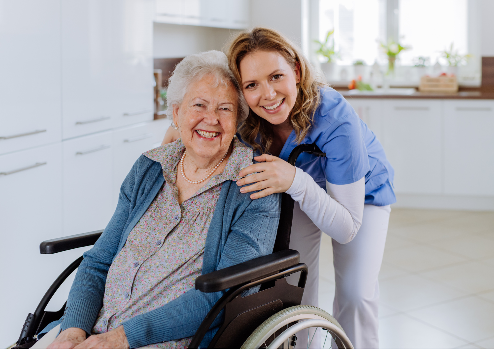 Starsza uśmiechnięta kobieta siedzi na wózku inwalidzkim a za nią stoi i obejmuje ją młoda uśmiechnięta kobieta ubrana jak pracownik medyczny. Znajdują się w domu, w kuchni.