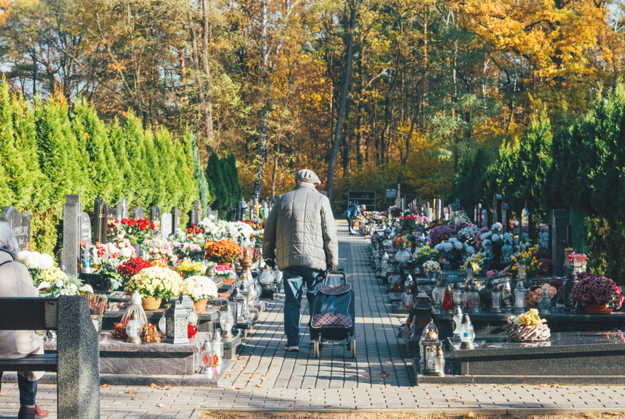 Starszy mężczyzna w alejce cmentarnej. Na froncie widać część ławki i siedzącą na niej kobietę.