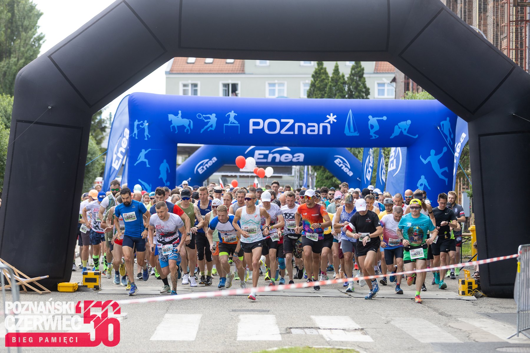 Start masowego biegu ulicznego »Poznański Czerwiec 56 - Bieg Pamięci«. Duża grupa biegaczy w kolorowych strojach sportowych przebiega pod wielką, czarną dmuchaną bramą startową. W tle widać kolejną, niebieską bramę z napisem »POZnan*« oraz logo sponsora »Enea«. Na pierwszym planie po lewej stronie znajduje się czerwono-białe logo wydarzenia z napisem »Bieg Pamięci«".