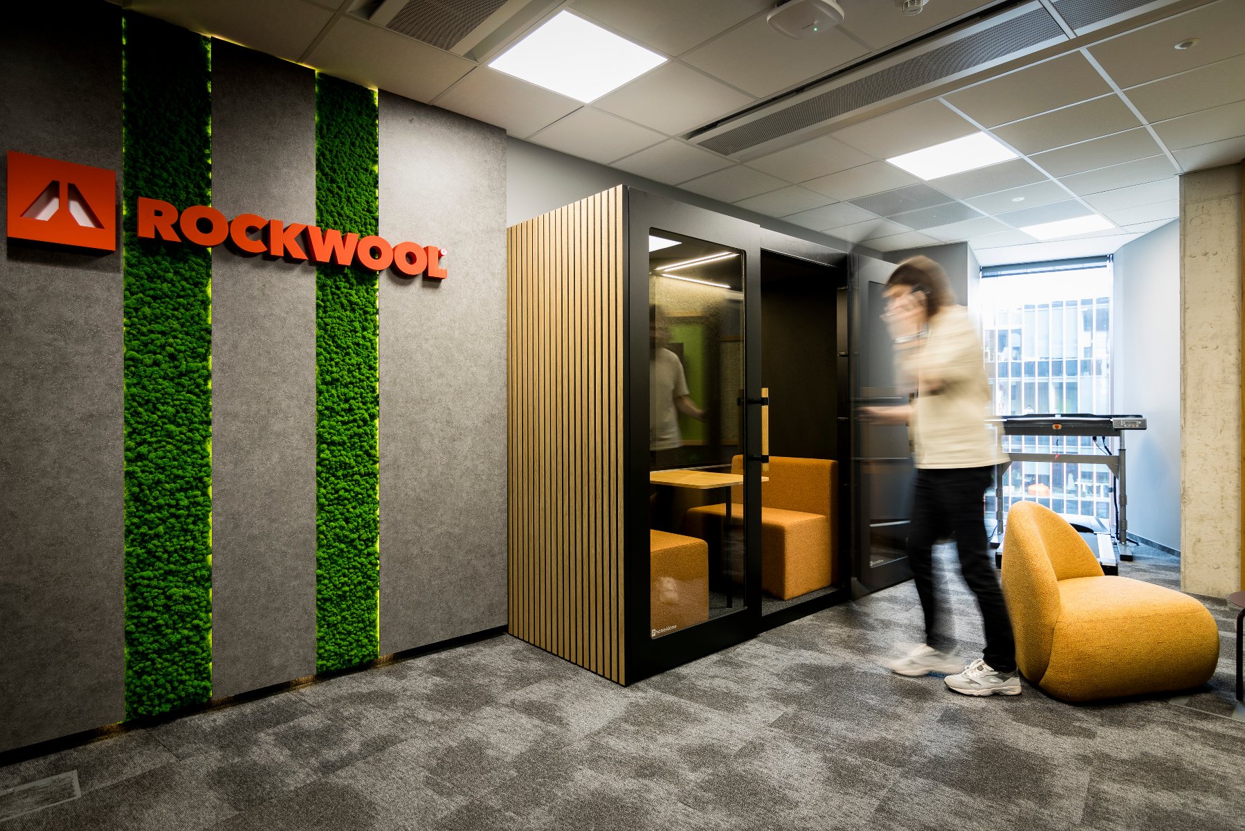 Strefa ciszy na korytarzu biurowym. Na ścianie z pasem zieleni wisi logo ROCKWOOL.