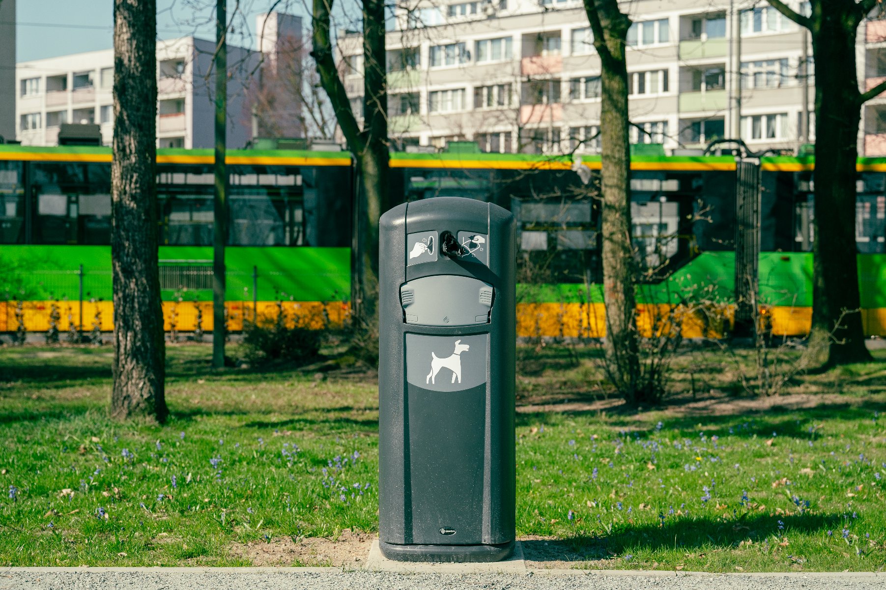 Szary kosz na psie odchody z piktogramem psa, stojący na tle zielono-żółtego tramwaju MPK Poznań i drzew. W tle widoczne są bloki mieszkalne. Kosz znajduje się na trawniku.