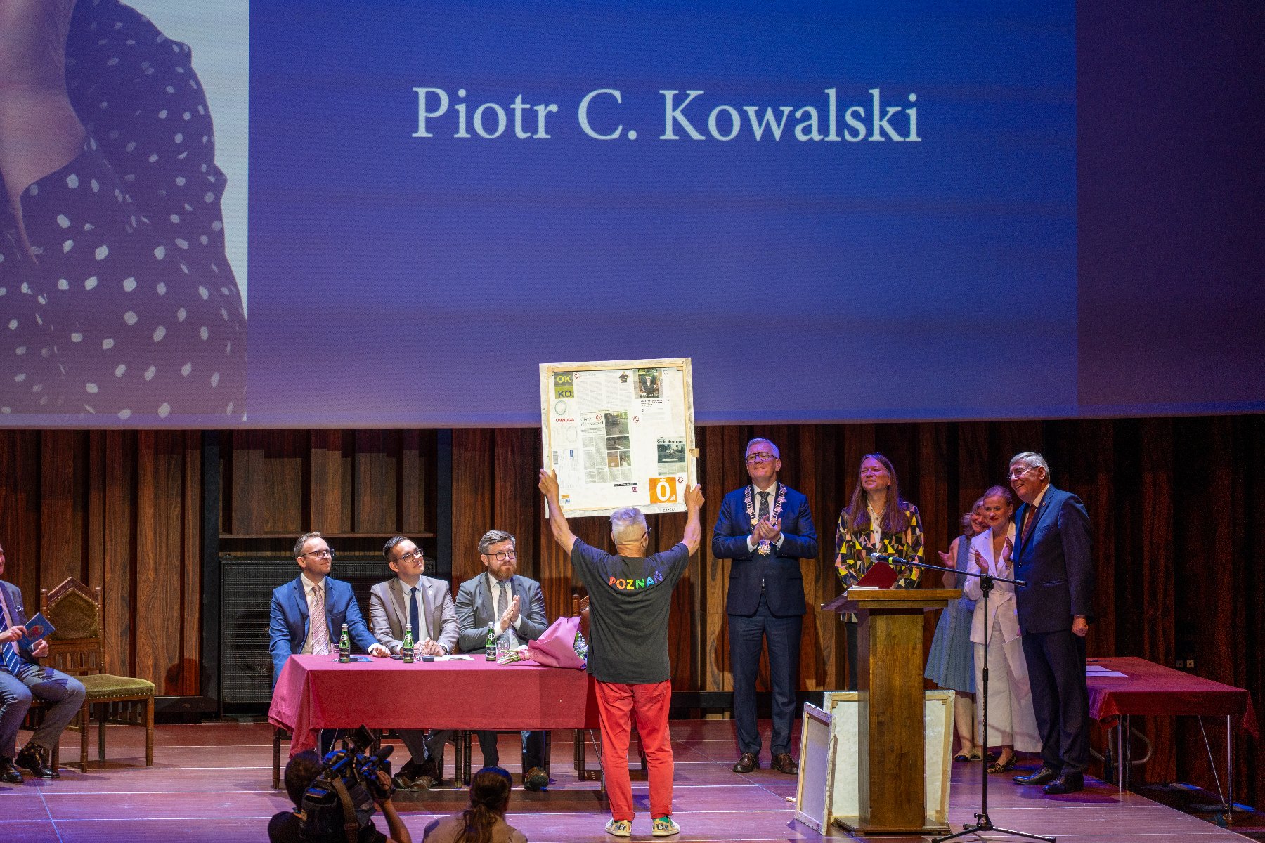Szerokie ujęcie sceny w auli. Na środku, tyłem do obiektywu, stoi mężczyzna w czarnej koszulce z kolorowym napisem "POZnan*" i czerwonych spodniach, unosząc oburącz duży, prostokątny obraz. Na scenie, za stołem przykrytym bordowym obrusem, siedzi trzech mężczyzn w garniturach. Po prawej stronie stoją kolejne osoby, w tym mężczyzna w oficjalnym stroju z łańcuchem na szyi. W tle widać duży ekran z napisem "Piotr C. Kowalski".