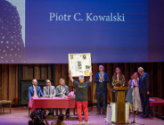 Szerokie ujęcie sceny w auli. Na środku, tyłem do obiektywu, stoi mężczyzna w czarnej koszulce z kolorowym napisem "POZnan*" i czerwonych spodniach, unosząc oburącz duży, prostokątny obraz. Na scenie, za stołem przykrytym bordowym obrusem, siedzi trzech mężczyzn w garniturach. Po prawej stronie stoją kolejne osoby, w tym mężczyzna w oficjalnym stroju z łańcuchem na szyi. W tle widać duży ekran z napisem "Piotr C. Kowalski".