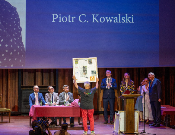 Szerokie ujęcie sceny w auli. Na środku, tyłem do obiektywu, stoi mężczyzna w czarnej koszulce z kolorowym napisem "POZnan*" i czerwonych spodniach, unosząc oburącz duży, prostokątny obraz. Na scenie, za stołem przykrytym bordowym obrusem, siedzi trzech mężczyzn w garniturach. Po prawej stronie stoją kolejne osoby, w tym mężczyzna w oficjalnym stroju z łańcuchem na szyi. W tle widać duży ekran z napisem "Piotr C. Kowalski".
