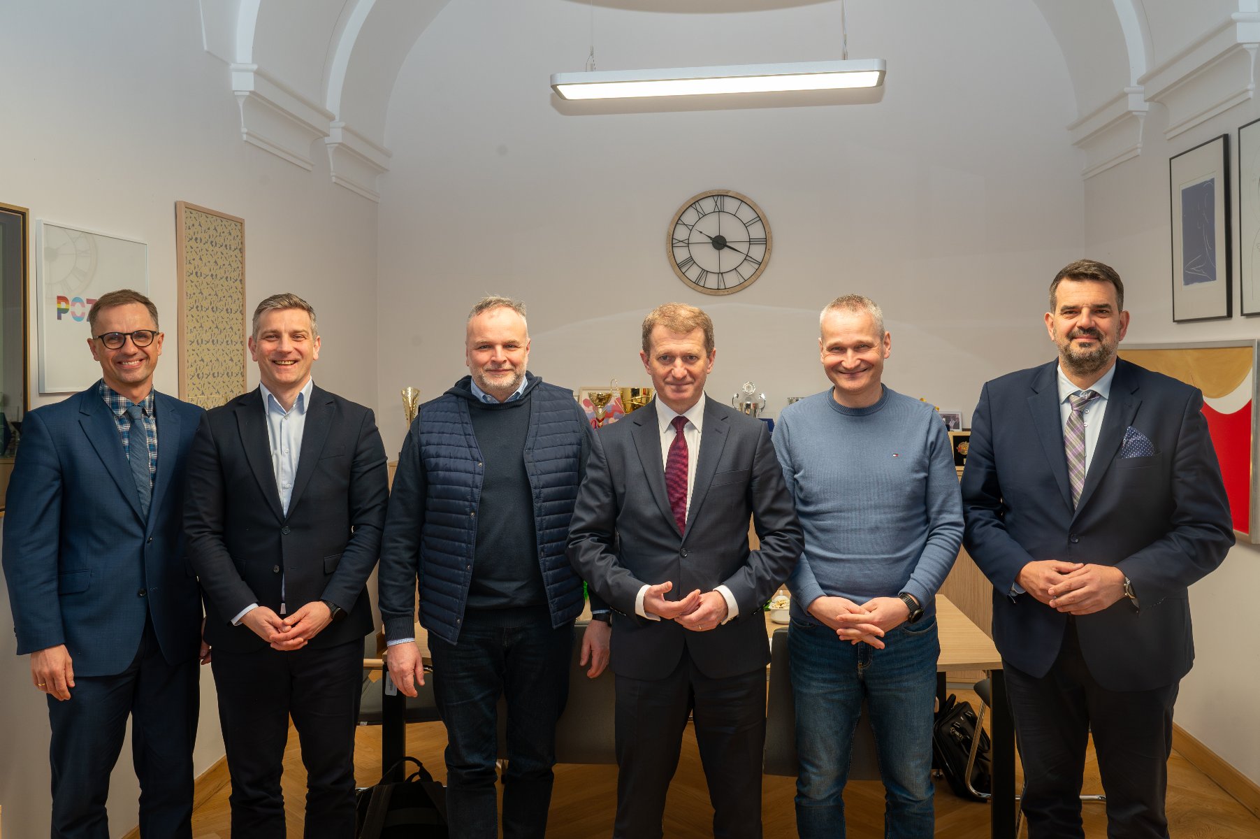 Sześciu mężczyzn stoi w eleganckim pokoju z wysokim sufitem i ozdobnymi łukami. Mężczyźni ubrani są w formalne i półformalne stroje, w tym garnitury, marynarki i swetry. Na ścianach pokoju widać zegar, trofea i nagrody na półkach i stołach, a także oprawione obrazy i zdjęcia.