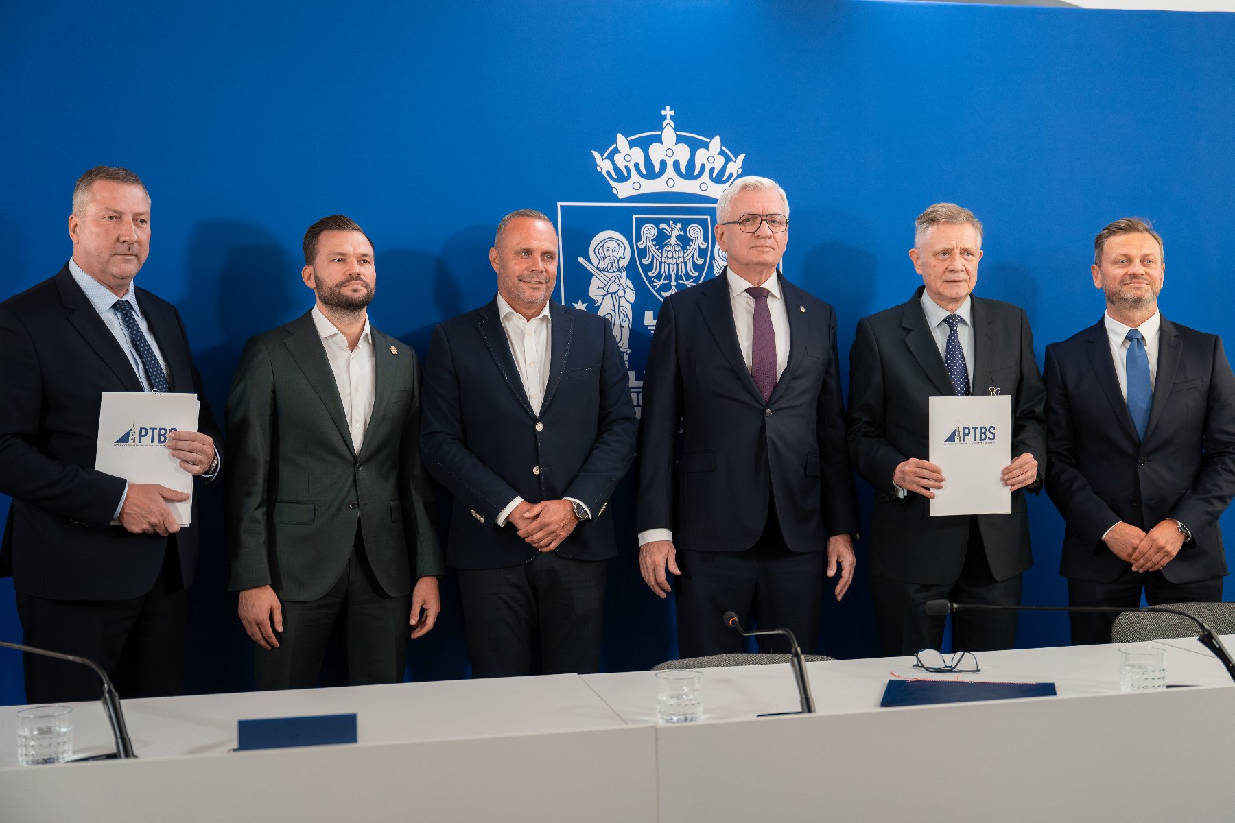 Sześciu mężczyzn w garniturach pozuje do pamiątkowego zdjęcia na tle niebieskiej ścianki z herbem Poznania, trzymając w rękach dokumenty z logo PTBS.