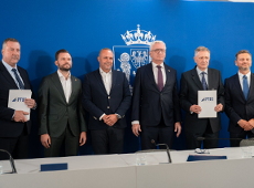 Sześciu mężczyzn w garniturach pozuje do pamiątkowego zdjęcia na tle niebieskiej ścianki z herbem Poznania, trzymając w rękach dokumenty z logo PTBS.