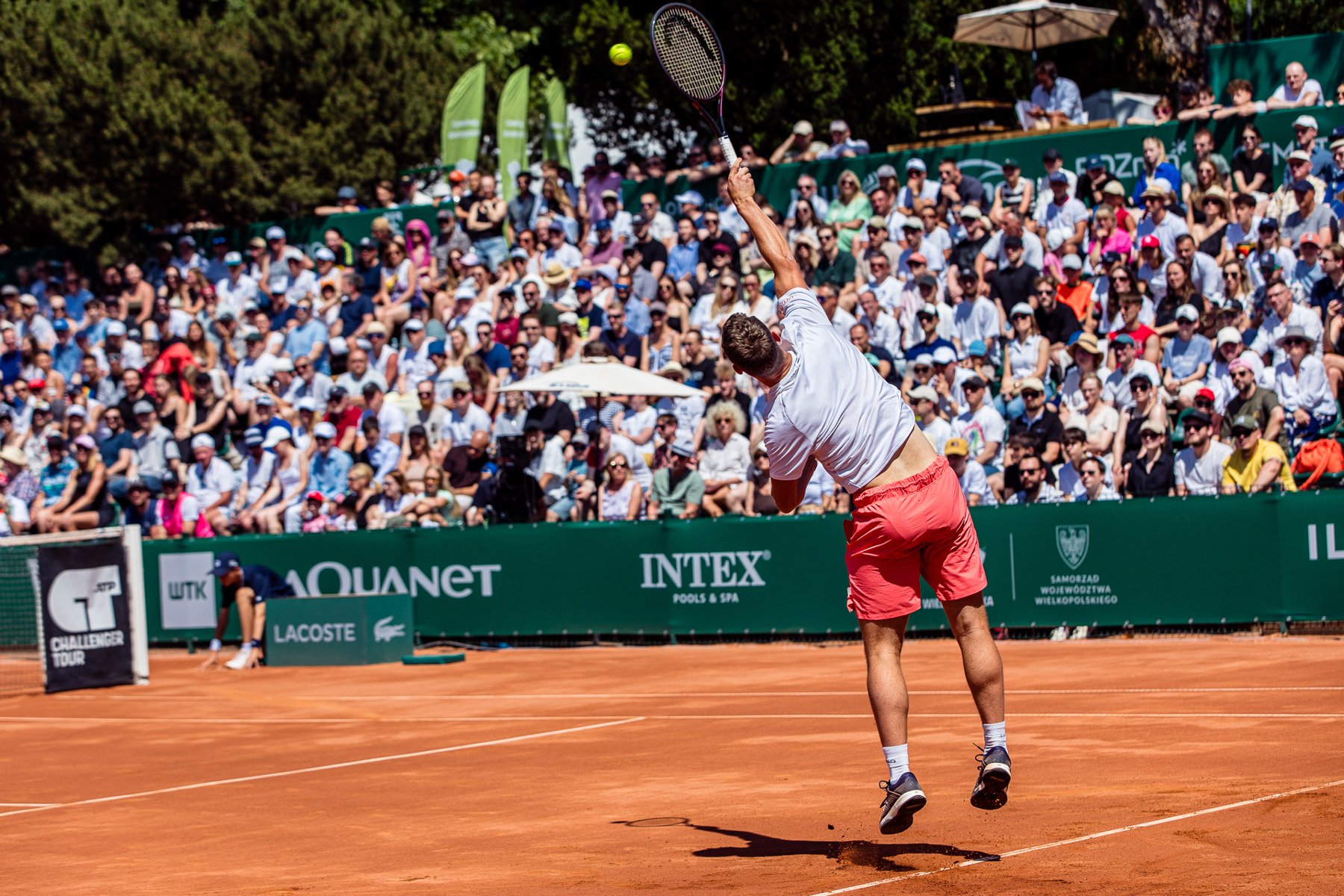 Tenisista w białej koszulce i koralowych spodenkach wykonuje serwis na korcie ziemnym. Zawodnik jest odwrócony tyłem, w wyskoku, z rakietą uniesioną wysoko nad głową w momencie uderzenia piłki. W tle widać gęsto zapełnione trybuny z publicznością.