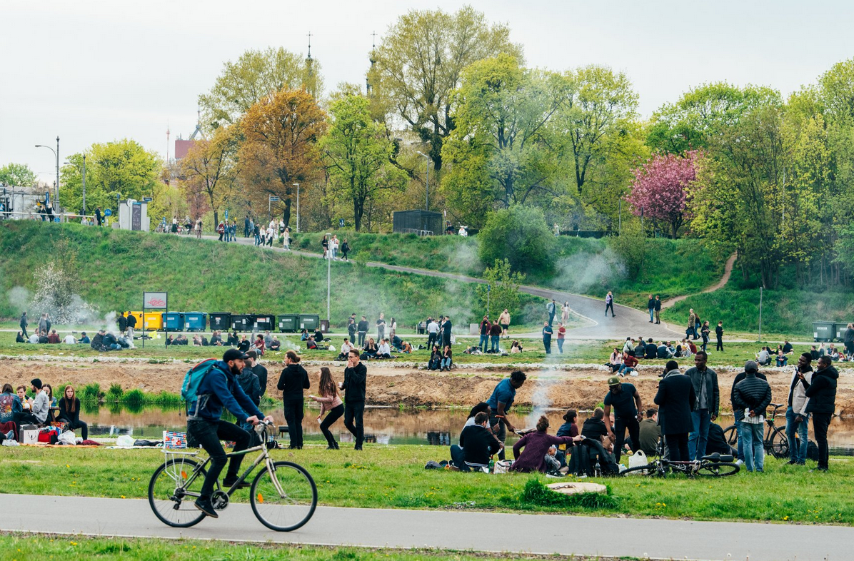 teren nad rzeką, na trawie sporo osób, siedzą lub stoją, grillują, rozmawiają, na dalszym planie wzniesienia terenu porośnięte trawą, na nich rosną drzewa
