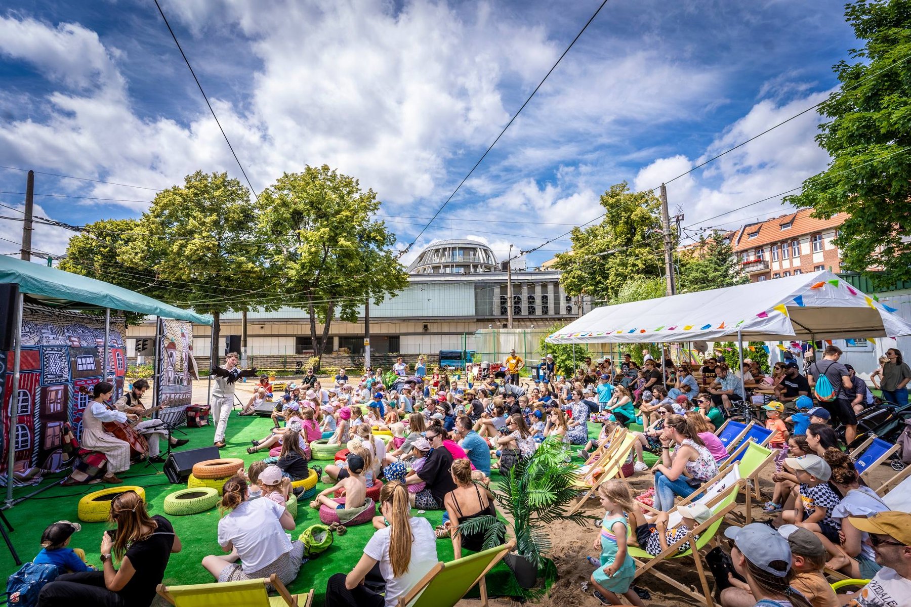 Tłum dzieci i dorosłych siedzi na plażowych leżakach przez sceną. Na scenie dwuosobowy zespół, kobieta gra na wiolonczeli a mężczyzna na gitarze. Za nimi dekoracje. W tle budynki i wysokie, zielone drzewa. Niebo jest blękitne i pokryte białymi chmurami.