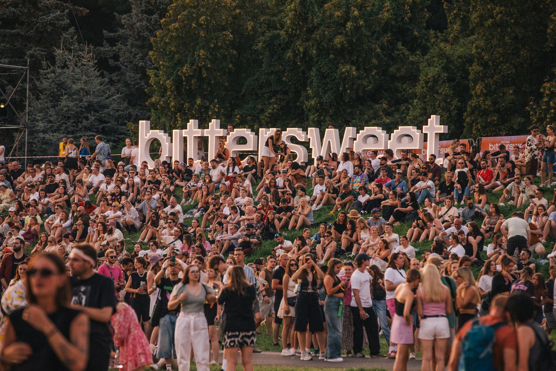 Tłum ludzi stoi na otwartym terenie przed sceną koncertową z dużym napisem "bittersweet". Na scenie widać ekrany i oświetlenie, a wydarzenie odbywa się w słoneczny dzień na tle drzew.