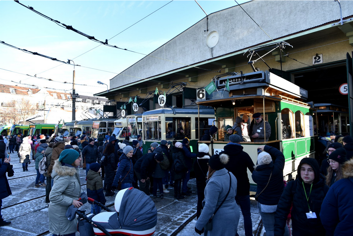 Tłum ludzi w zimowych ubraniach ogląda zabytkowe i współczesne tramwaje ustawione przed dużą zajezdnią. Kilka tramwajów ma numery liniowe na tablicach, a w jednym z nich widać obsługę w środku rozmawiającą z odwiedzającymi. Na ziemi leży cienka warstwa śniegu.