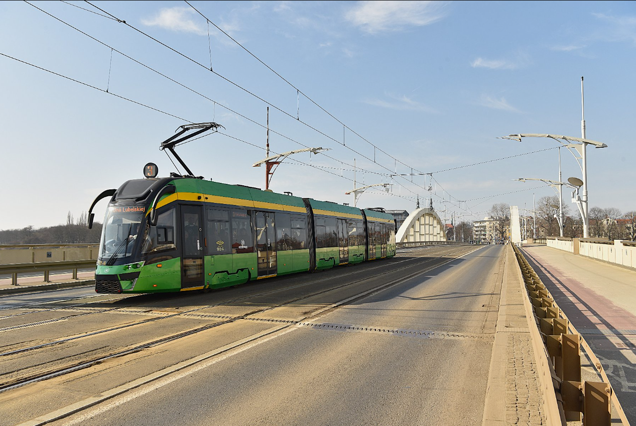 Tramwaj jadący po torach w miejskim otoczeniu. W tle widoczny most i drzewa.