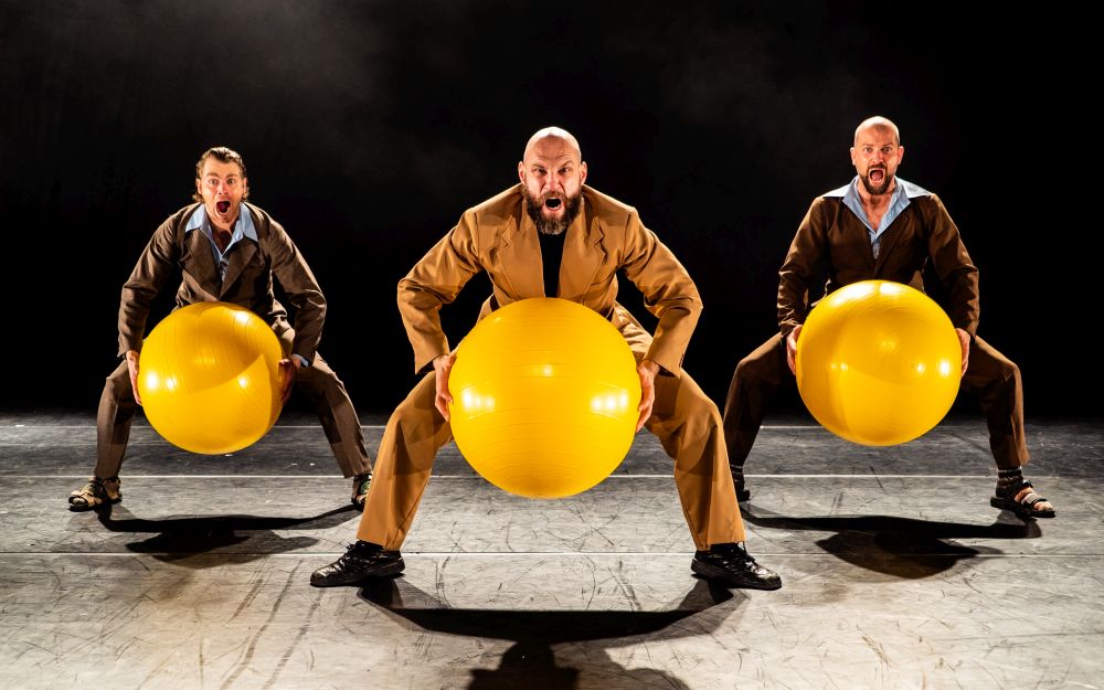Trzech mężczyzn kuca z wielkimi, żółtymi piłkami.