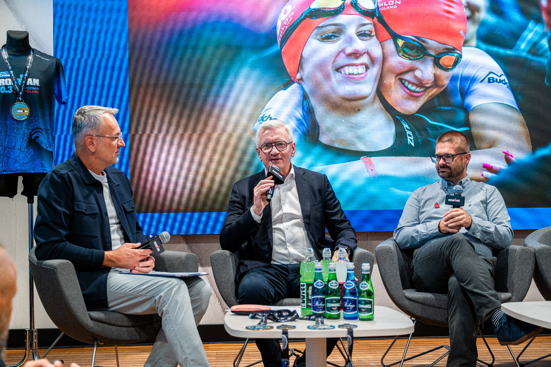 Trzech mężczyzn siedzących na fotelach podczas panelu dyskusyjnego, z mężczyzną w okularach przemawiającym do mikrofonu w centrum, na tle ekranu ze zdjęciem pływaczek.