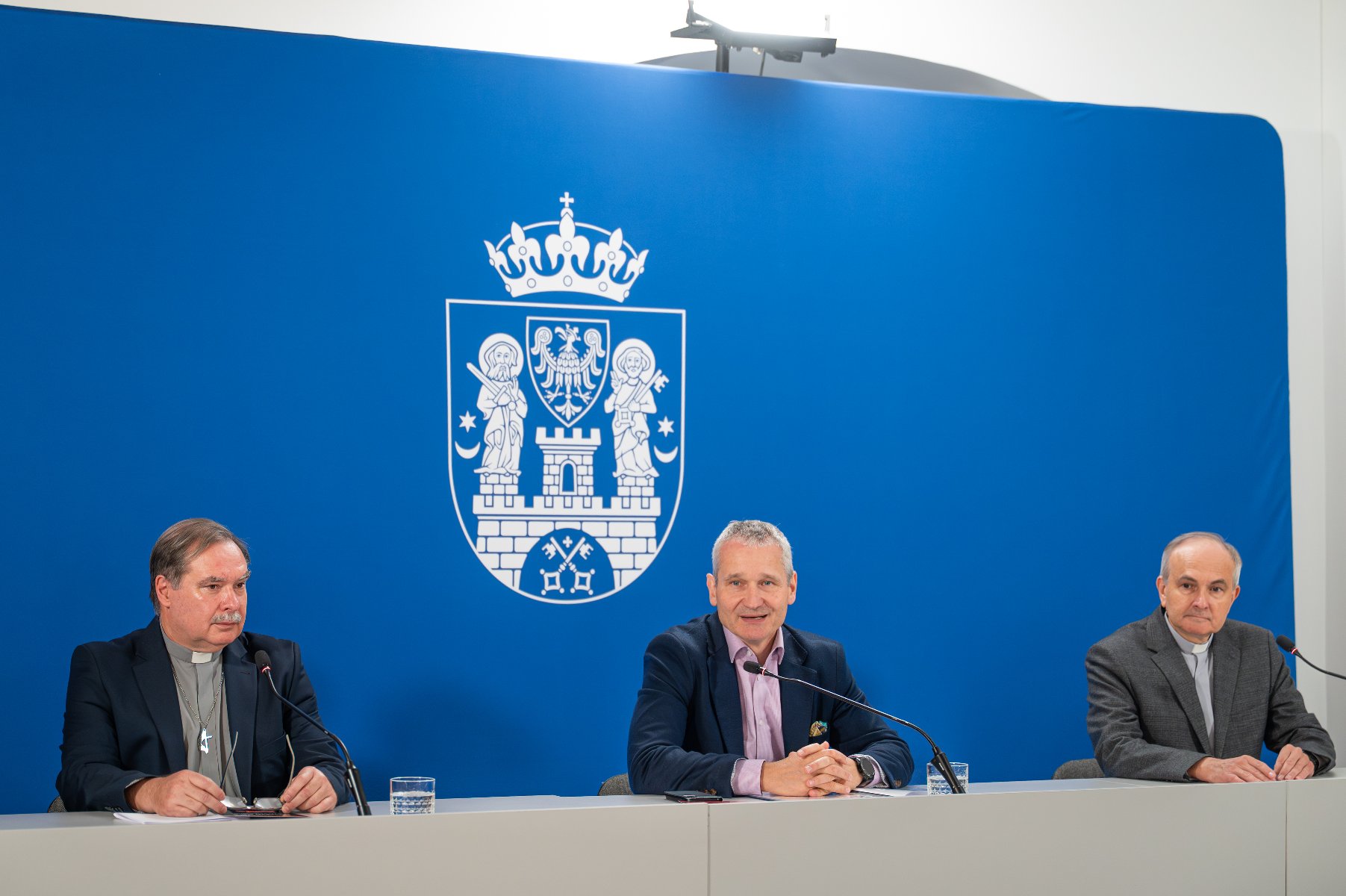 Trzej mężczyźni siedzą za długim, szarym stołem podczas konferencji prasowej lub spotkania. W tle wisi duże, niebieskie tło z herbem Poznania w kolorze białym. Mężczyzna po lewej, ubrany w sutannę i krzyż na łańcuszku, jest duchownym. Mężczyzna pośrodku jest uśmiechnięty i ma na sobie niebieską marynarkę, a trzeci mężczyzna siedzi po prawej, ubrany w szary garnitur.