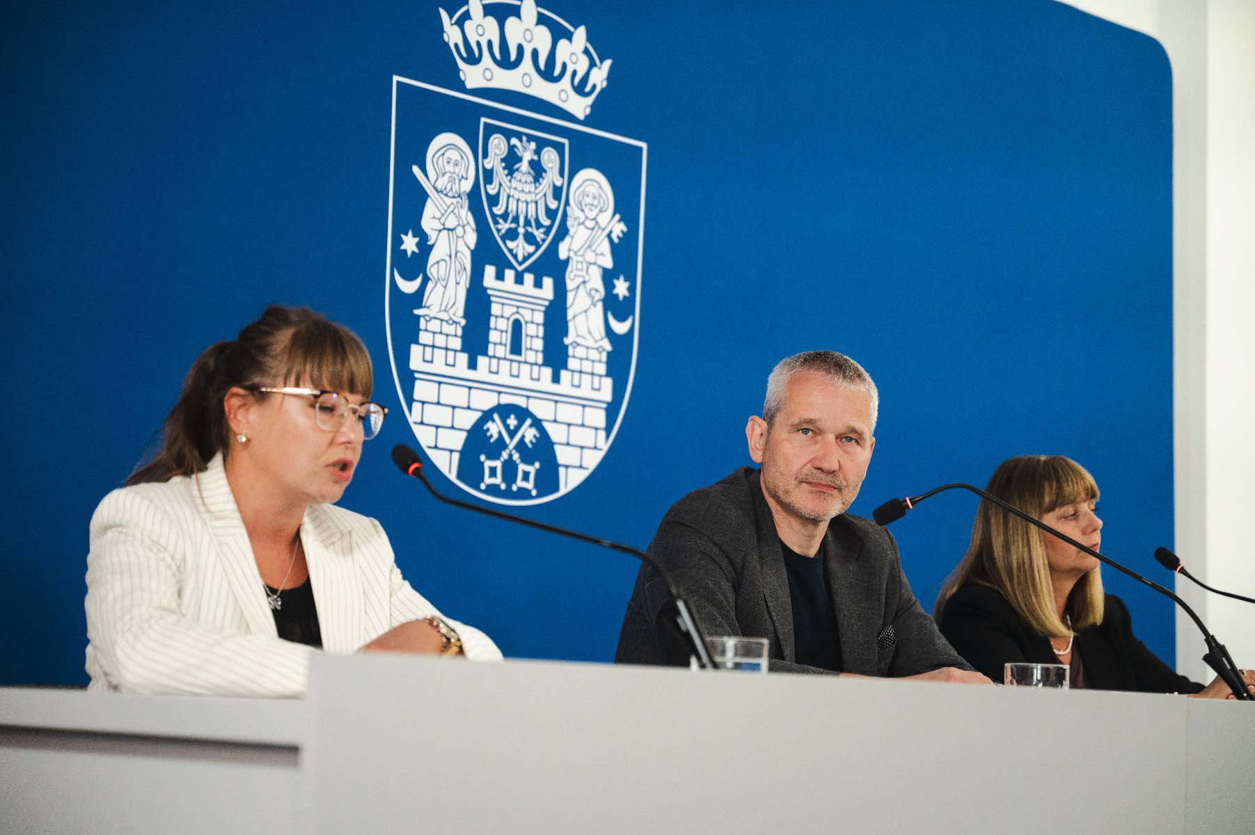 trzy osoby na konferencji prasowej, w tle niebieska ściana i herb Poznania