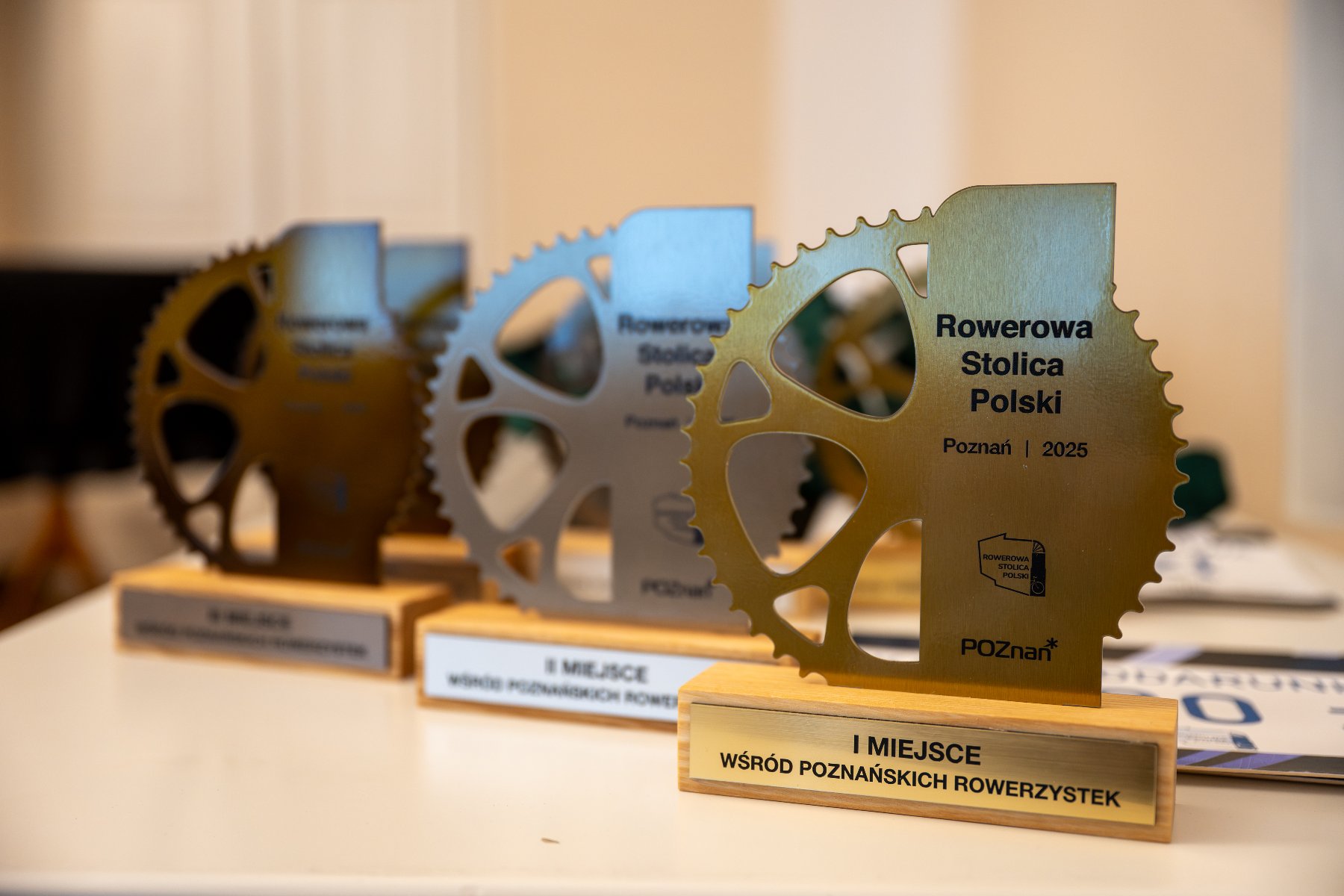 Trzy statuetki w kształcie rowerowych zębatek ustawione na białym stole. Na pierwszym planie złota nagroda z napisem: "Rowerowa Stolica Polski, Poznań 2025, I miejsce wśród poznańskich rowerzystek". W tle widoczne są kolejne trofea w srebrnym kolorze.