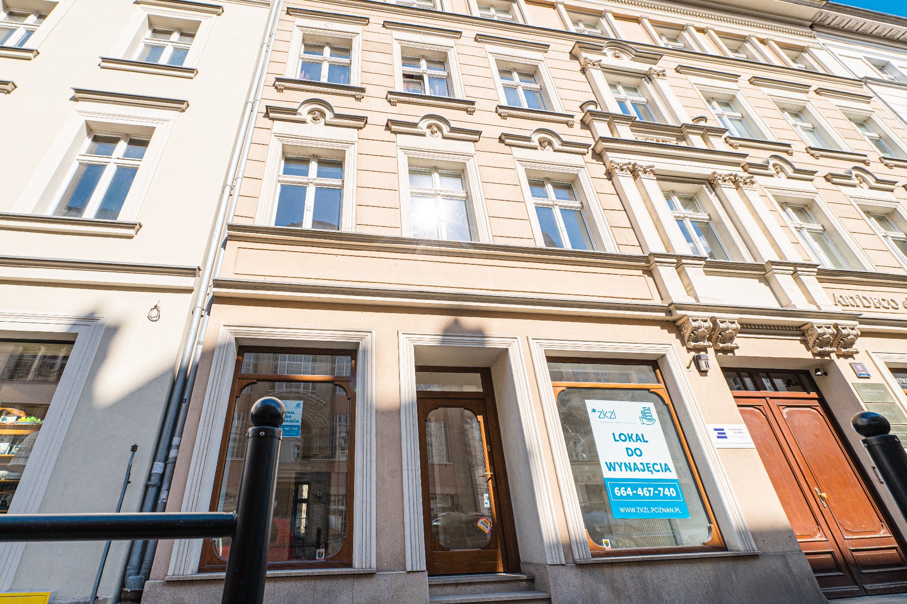 Trzypiętrowa beżowa kamienica. Na parterze znajdują się trzy duże okna wystawowe oraz drewniane drzwi wejściowe. W prawym oknie plakat z napisem "Lokal do wynajęcia" oraz numerem telefonu i adresem strony internetowej.