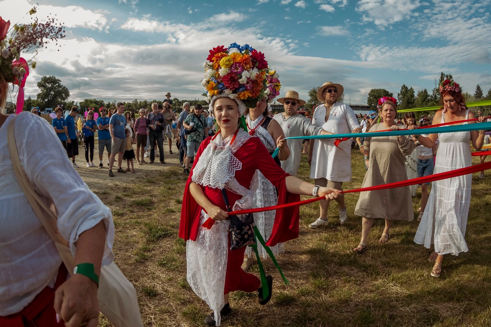 Uczestnicy barwnego korowodu plenerowego, idący po trawiasstym terenie nad rzeką. Na pierwszym planie wyróżnia się kobieta w czerwonym stroju z białym, koronkowym kołnierzem, niosąca na głowie potężne, wysokie nakrycie ozdobione licznymi, kolorowymi kwiatami. W dłoniach trzyma długie, czerwone i zielone wstęgi, które ciągną się za nią, trzymane przez kolejne osoby w białych i beżowych ubraniach, z których część ma na sobie słomkowe kapelusze.