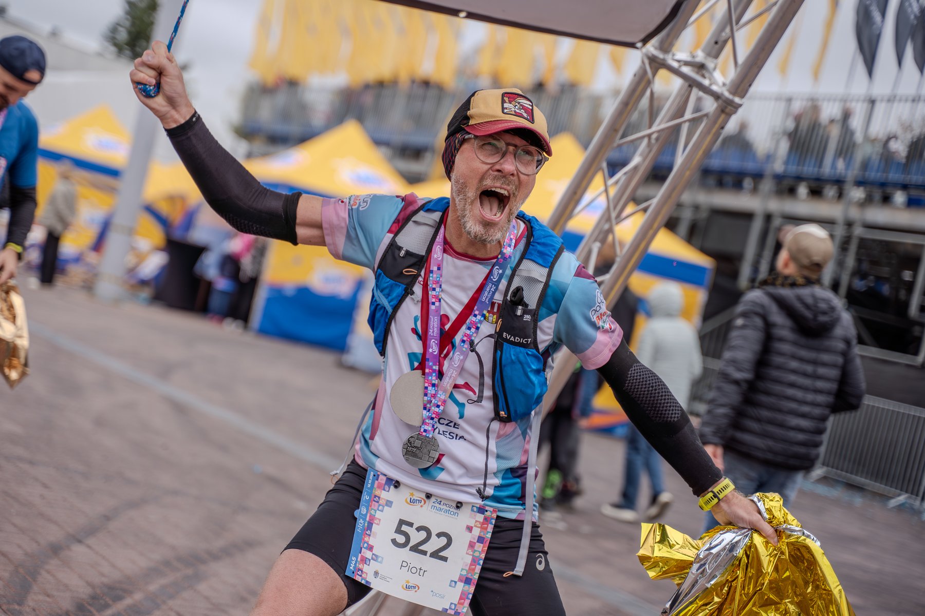 Uczestnik biegu z numerem startowym 522 na mecie maratonu. Na szyi ma kilka medali, na plecach kamizelkę do biegania, a w dłoniach trzyma folię termiczną. W tle widoczne są żółto‑niebieskie namioty, metalowa konstrukcja oraz inne osoby znajdujące się w strefie mety.