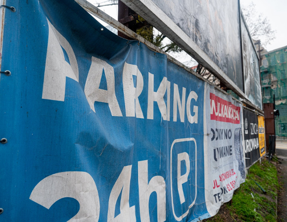 Ujęcie z bliska na pomarszczony, niebieski baner reklamowy, na którym białym kolorem widnieje duży napis w cudzysłowie: "PARKING 24h" i symbol litery P.