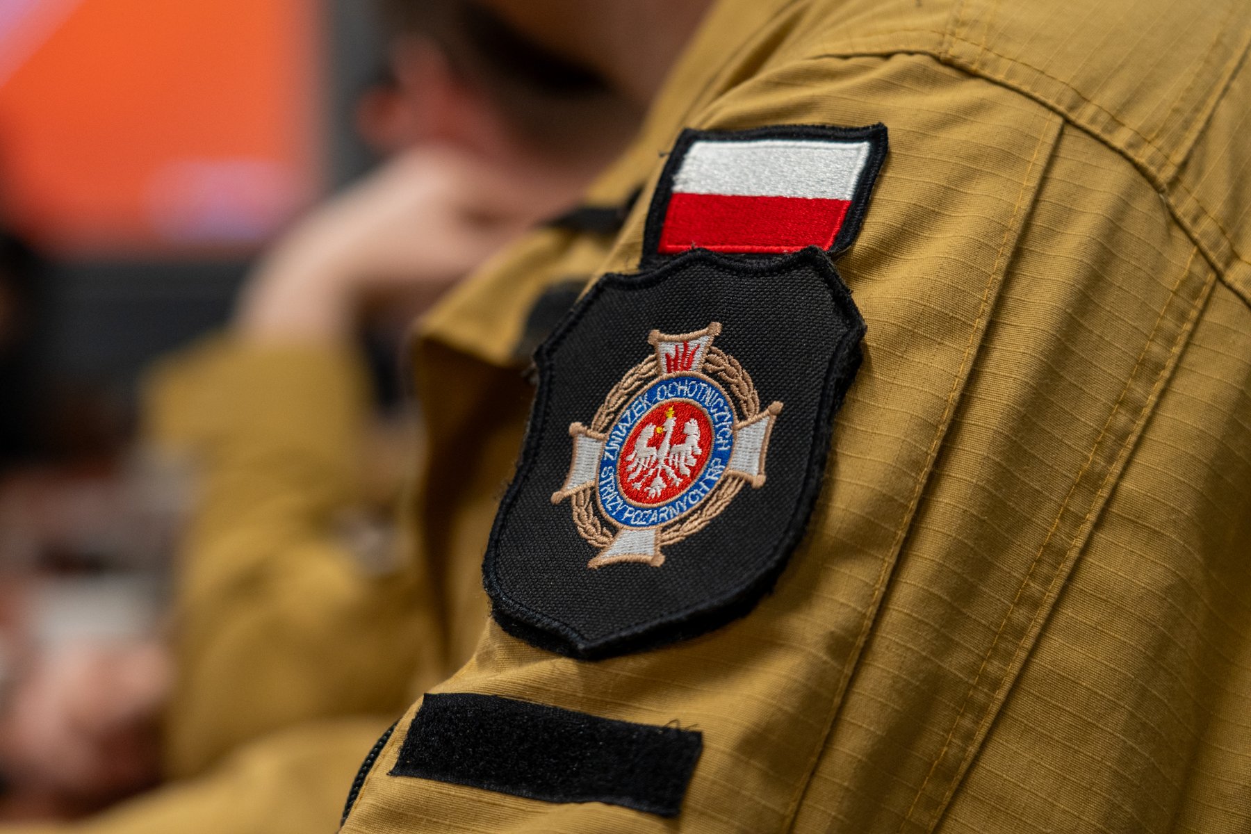 Ujęcie z bliska na ramię w beżowym mundurze. Na ramieniu widoczna jest naszywka z polską flagą oraz naszywka ze złotymi i czerwonymi elementami oraz herbem, zawierająca napis w cudzysłowie: "ZWIĄZEK OCHOTNICZYCH STRAŻY POŻARNYCH".