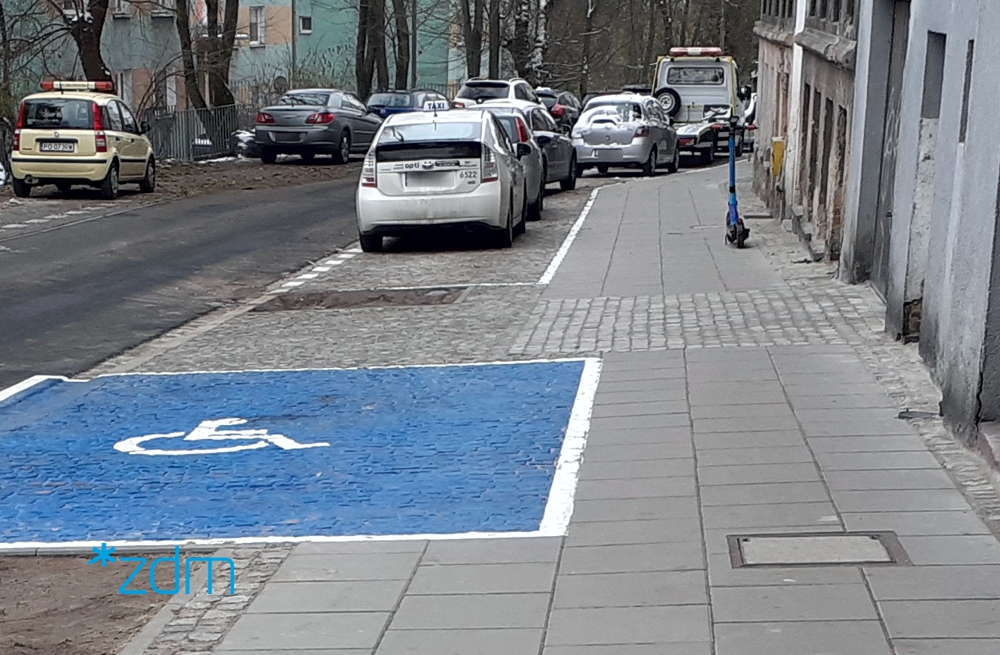 ulica, przy chodnikach zaparkowane samochodwy, widoczna wolna koperta dla samochodów osób niepełnosprawnych