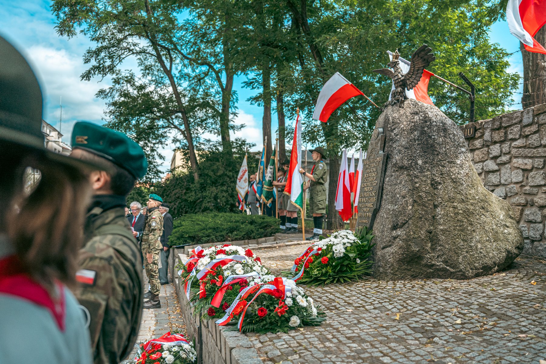 Uroczystość patriotyczna na zewnątrz. Na pierwszym planie widoczne tyły głów żołnierzy w mundurach, z emblematami na ramieniu. Po prawej duży kamień z rzeźbą orła na szczycie i wmurowaną tablicą. U podnóża kamienia oraz wzdłuż kamiennego murku leżą biało-czerwone wieńce. W tle widoczne poczty sztandarowe i polskie flagi.