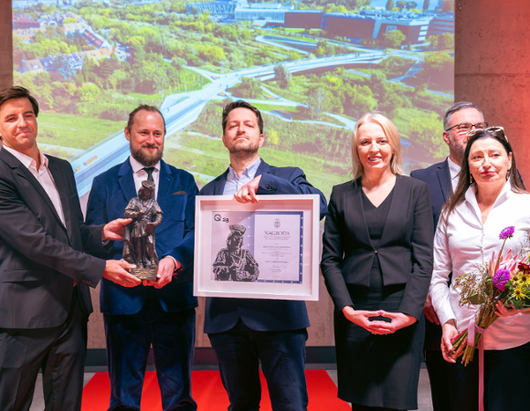 Uroczystość wręczenia nagrody architektonicznej z udziałem laureatów i przedstawicieli władz na tle prezentacji multimedialnej przedstawiającej panoramę inwestycji miejskiej.