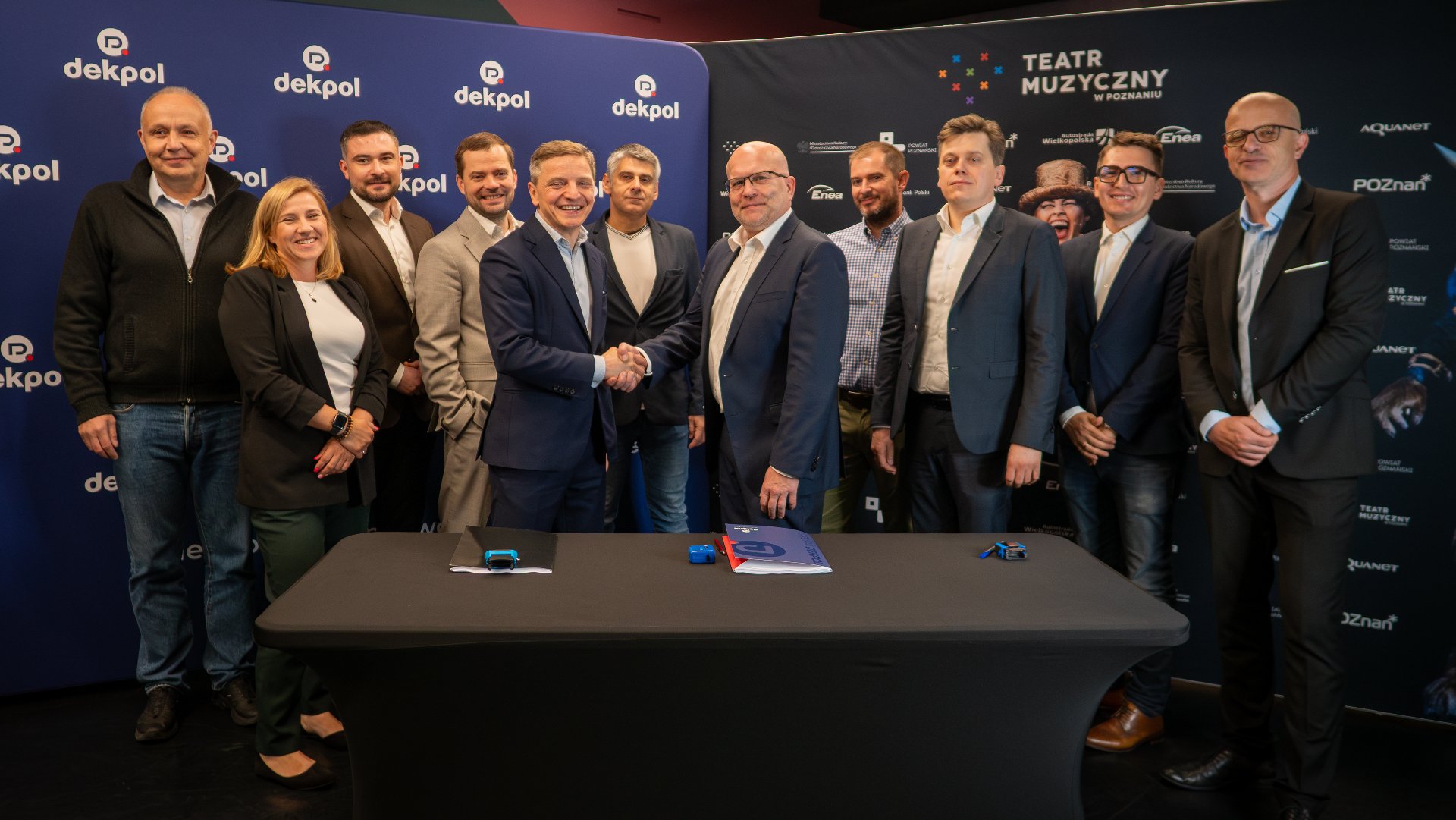 Uścisk dłoni dwóch panów. W tle grupa ludzi ubranych elegancko. Świętują podpisanie umowy.