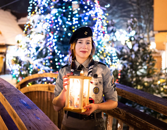 Uśmiechnięta młoda harcerka w szarym mundurze, ciemnym berecie i z odznakami, stoi na drewnianym, ośnieżonym mostku i trzyma w dłoniach lampion ze Światłem Betlejemskim. W tle widoczna jest duża, udekorowana choinka świąteczna z licznymi niebieskimi i białymi lampkami.