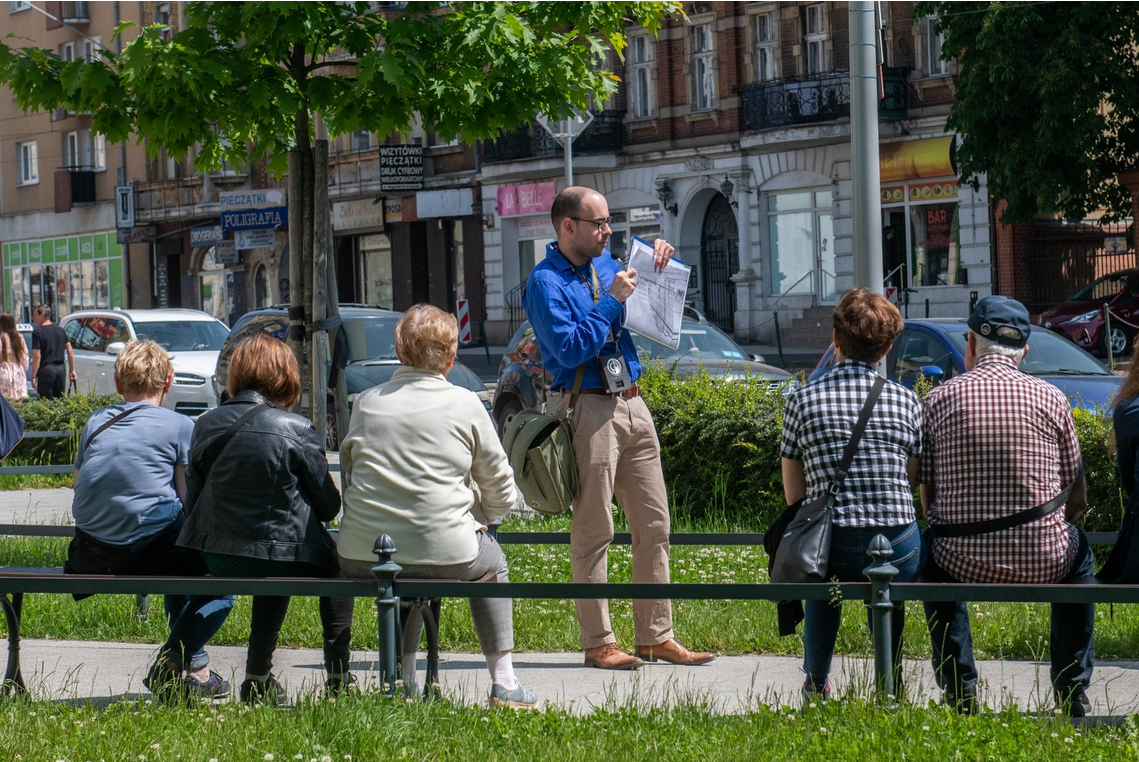 W centralnej części grafiki przewodnik trzymający mapę. Wokół grupa słuchaczy. W tle ulica i budynki poznańkich kamienic.