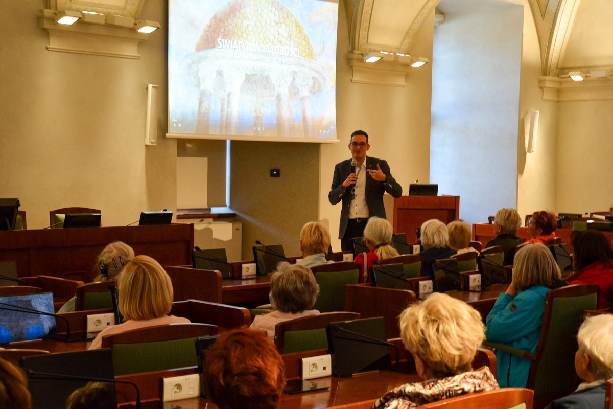 w dużej sali grupa osób siedzi przy stanowiskach z mikrofonami przed sto nimi mężczyzna z mikrofonem i mówi do nich