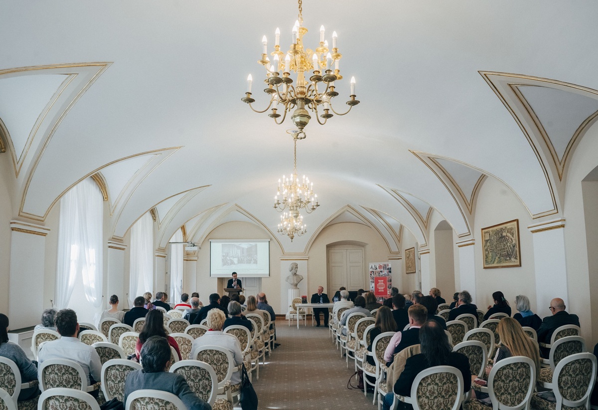 W reprezentacyjnej zabytkowej sali odbywa się spotkanie na krzesłach ustawiony w rzędach siedzi publiczność, przed nimi stół prezydialny, przy którym siedzą dwie osoby, obok mównica przy której stoi jedna osoba - grafika artykułu
