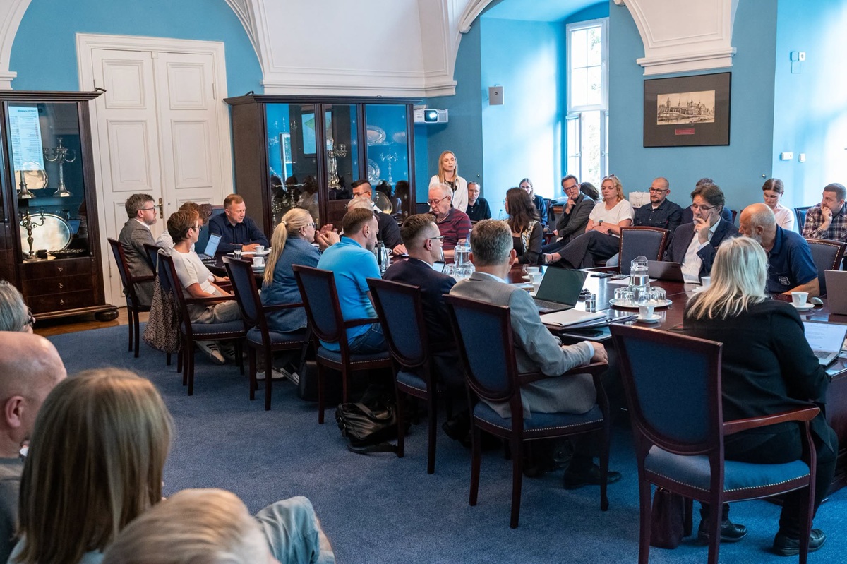 W sali obradują radne i radni przy stole konferencyjnym, wzdłuż ścian na krzesłach siedzą inne osoby