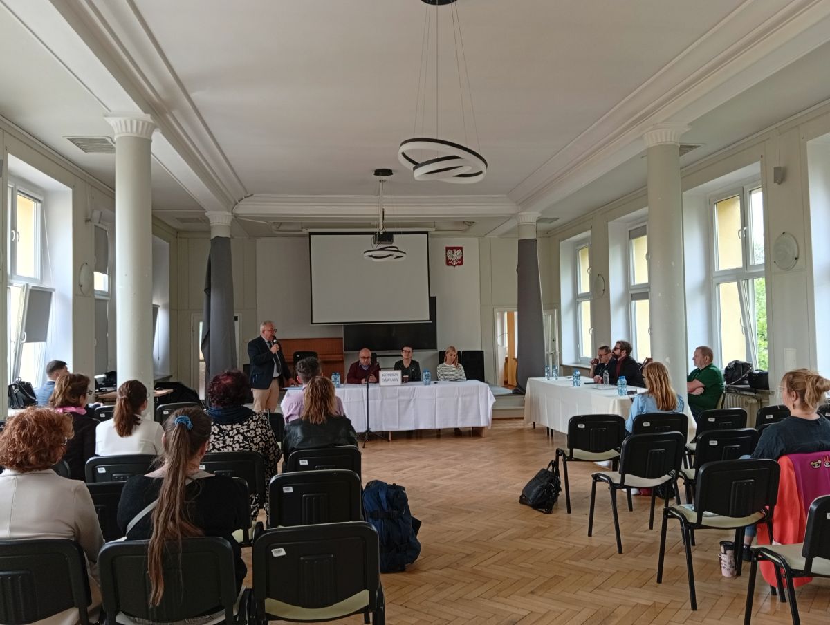 w sali obraduje większa liczba osób, trzy osoby siedzą za stołem prezydialnym, kilka osób za stołem obok, jedna osoba stoi, pozostałe siedzą na krzesłach ustawionych w rzędach przed stołem prezydialnym