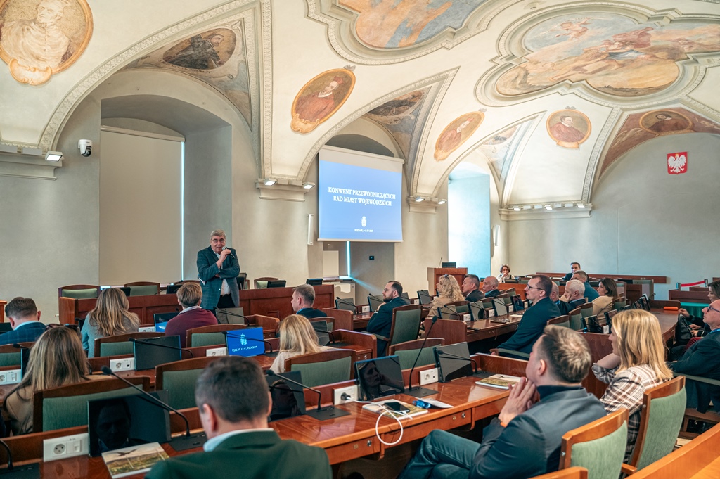 w sali sesyjnej przy stanowiskach z monitoriami i mikrofonami siedzą osoby, przed nimi stoi jedna osoba - mężczyzna trzymający mikrofon, sklepienie sali zdobią kolorowe, zabytkowe freski
