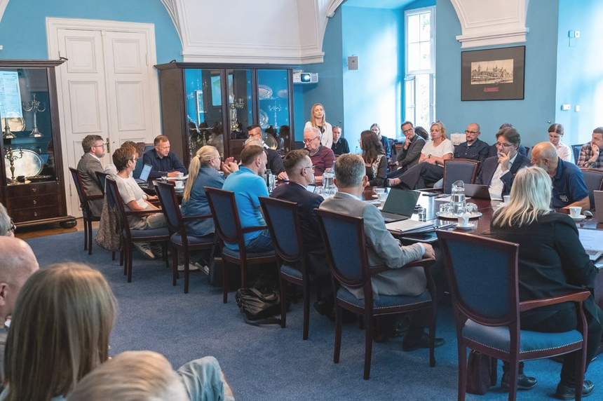 w sali za stołem konferencyjnym obraduje grupa ci najmniej kilkunastu osób, inne osoby siedzą na krzesłach wzdłuż ściany