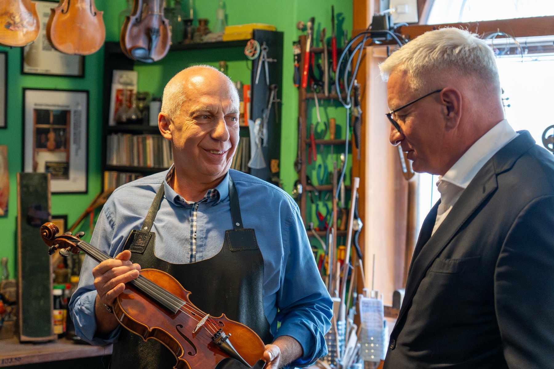 W warsztacie lutniczym dwóch mężczyzn rozmawia. Mężczyzna po lewej, ubrany w niebieską koszulę i czarny fartuch, uśmiecha się i trzyma w rękach skrzypce. Mężczyzna po prawej jest w ciemnym garniturze i okularach. W tle widać zieloną ścianę i liczne narzędzia lutnicze.
