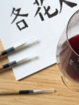 Kartka papieru a na niej chińska kaligrafia, obok kieliszek z czerwonym napojem.